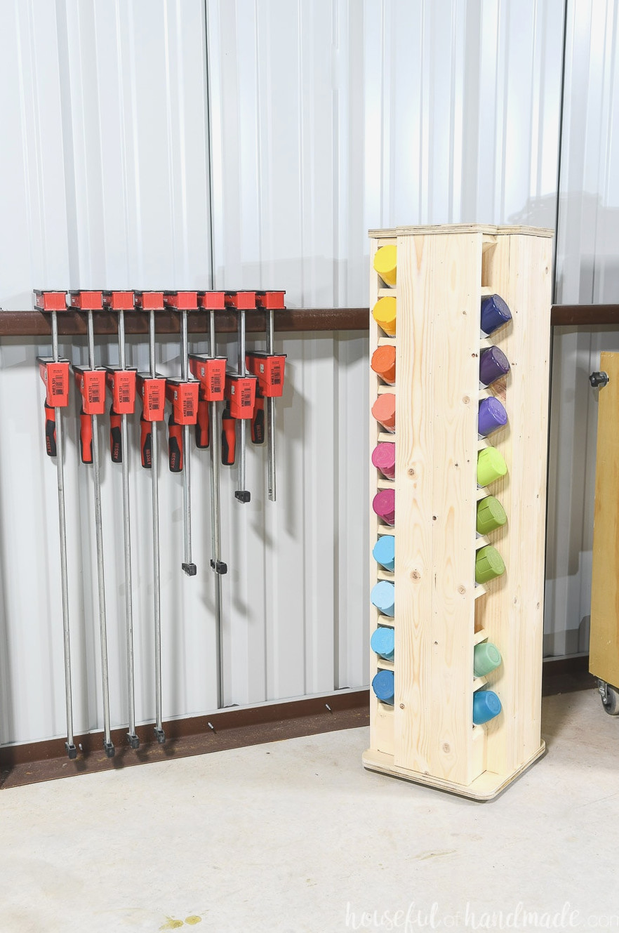 Compact rotating spray paint storage cabinet with cans arranged in a rainbow in a corner beside bar clamps in a workshop.