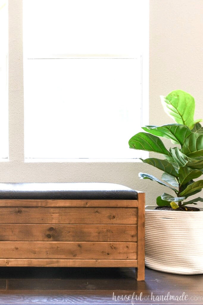 A brown-stained wooden storage bench with one side upholstered in a black faux-leather cushion, positioned beside a plant in a fabric basket.