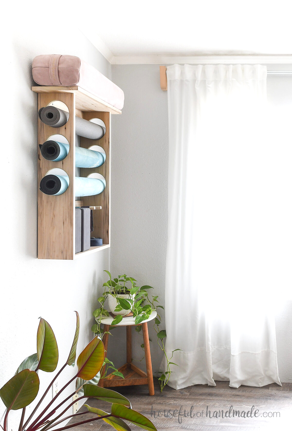 Wood DIY yoga mat holder on the wall with 3 yoga mats inside and plants on the floor by it.