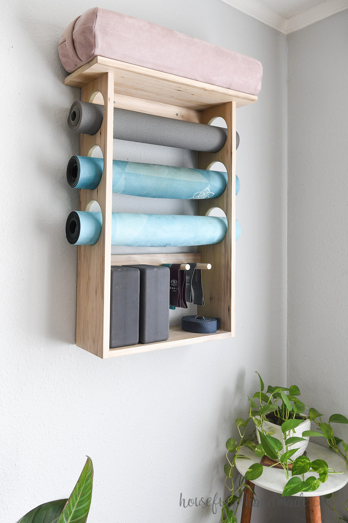 Three yoga mats in a wall mounted holder with a bolster on the top and blocks below.