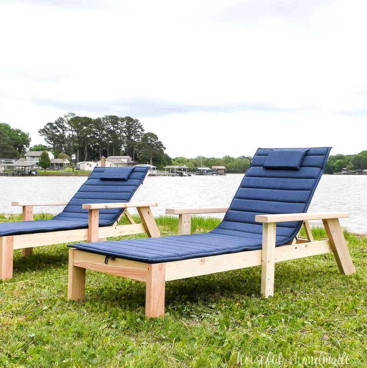 Two wooden pool chaise lounge chairs sitting on the grass beside a lake.