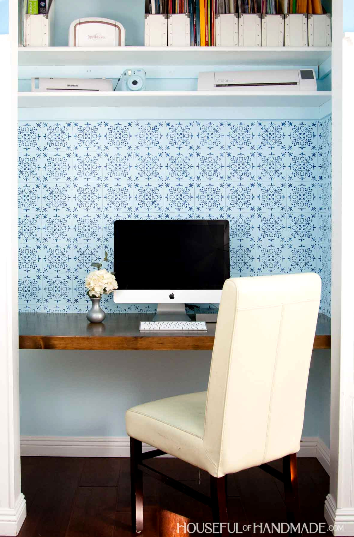 DIY closet desk featuring a wooden desktop, blue patterned wallpaper, an Apple computer, and an office chair.