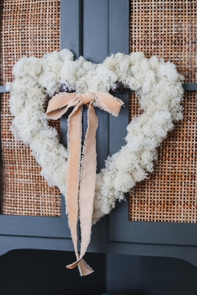 a white reindeer moss heart shaped wreath with a blush colored velvet ribbon hanging down